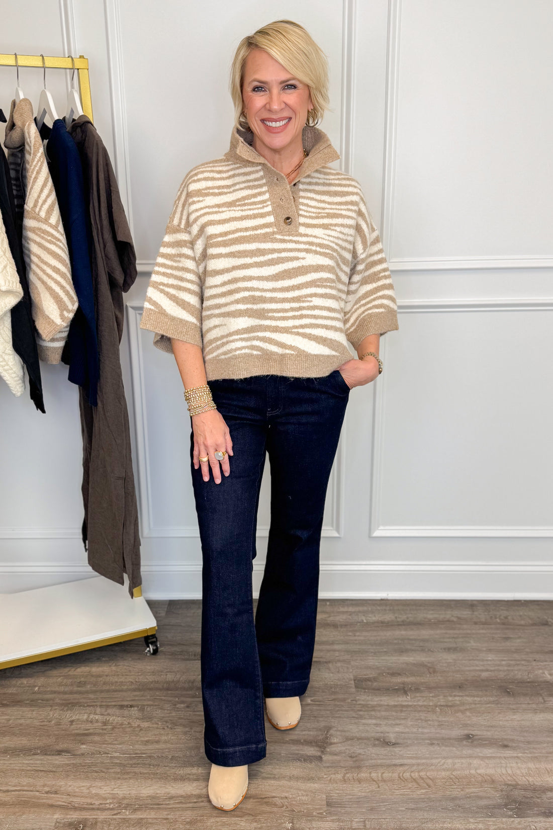 A woman modeling a beige and cream striped knit polo sweater with dark blue flare jeans and beige heels, standing in a boutique setting with a clothing rack visible on the left.