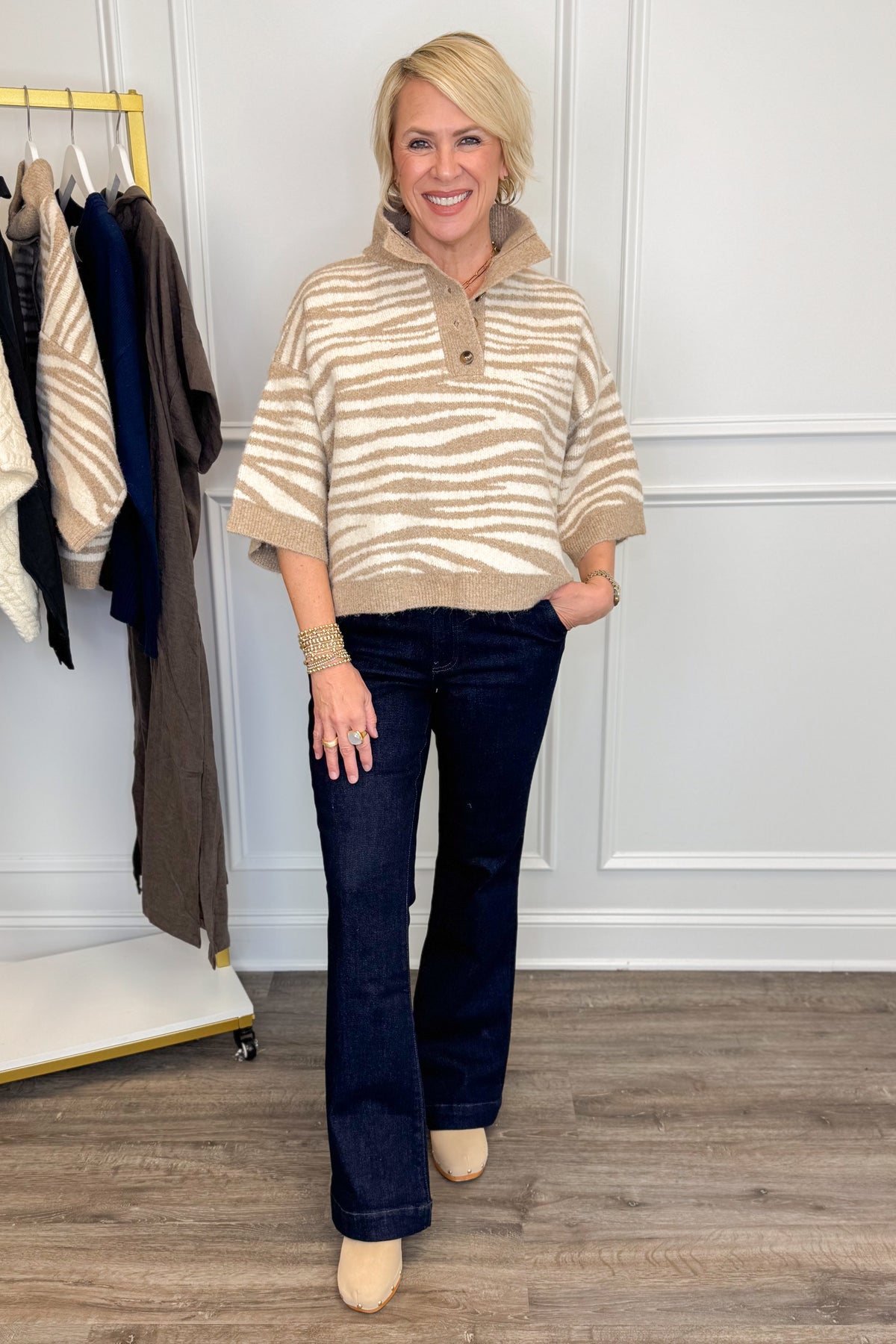 A woman modeling a beige and cream striped knit polo sweater with dark blue flare jeans and beige heels, standing in a boutique setting with a clothing rack visible on the left.