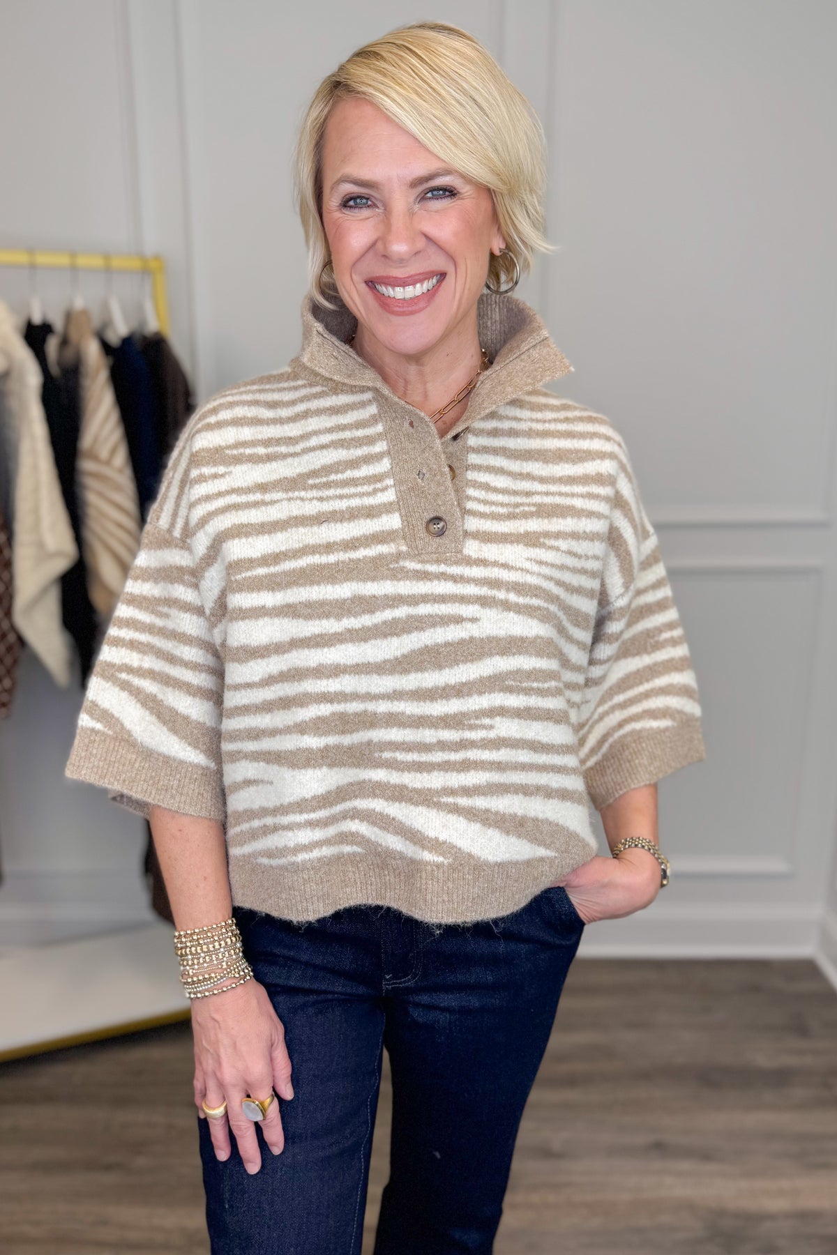 A smiling blonde woman models a beige and off-white horizontally striped knit polo sweater with a buttoned collar, paired with dark blue jeans in a boutique setting.