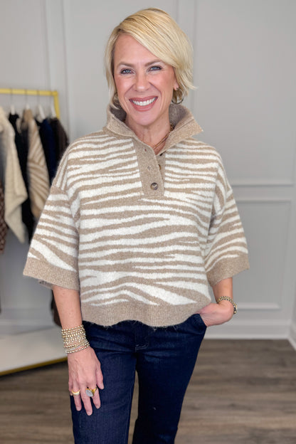 A smiling blonde woman models a beige and off-white horizontally striped knit polo sweater with a buttoned collar, paired with dark blue jeans in a boutique setting.
