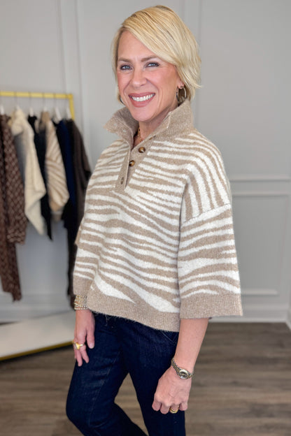A smiling blonde woman wearing a beige and off-white striped knit mock-neck sweater with a short button placket, paired with dark jeans, standing in a boutique setting.