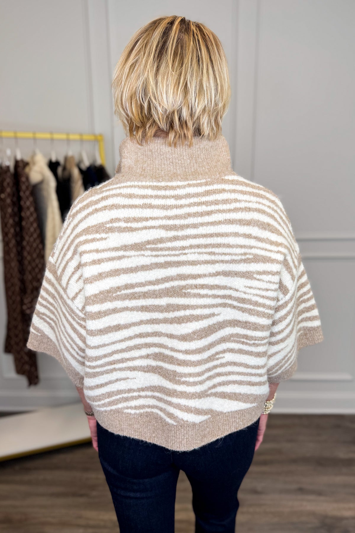 A woman viewed from behind wearing a beige and white wavy striped knit poncho sweater, paired with dark blue jeans, in a boutique setting.