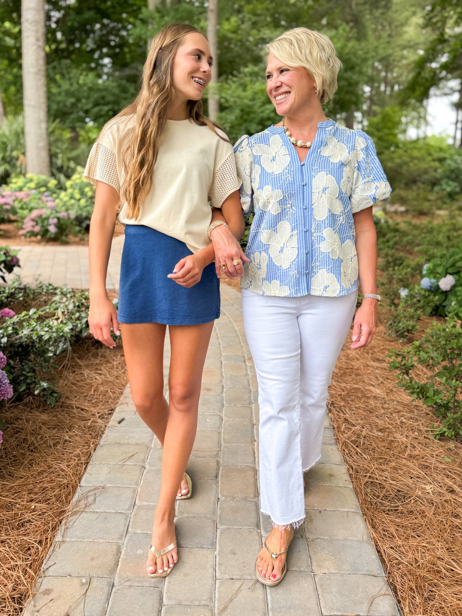 Front view of woman wearing Swagger Boutique’s Indigo Gail Jersey Denim Mini Skirt by Z Supply with Island Coconut Banyan Top by Z Supply and woman wearing jeans & floral top walking down a walkway outside.