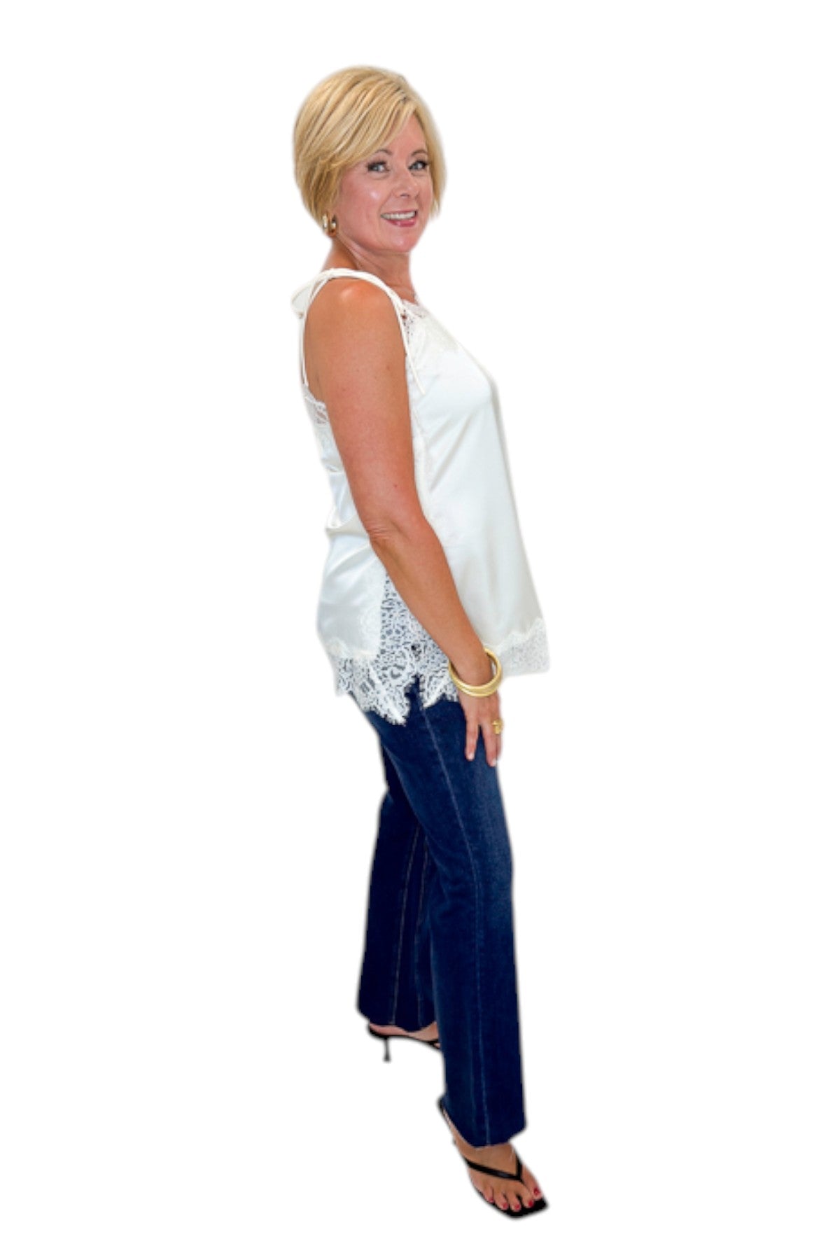 Side view of woman wearing Swagger Boutique’s Ivory Satin Lace Trim Tank Top by Entro with jeans against white studio background.