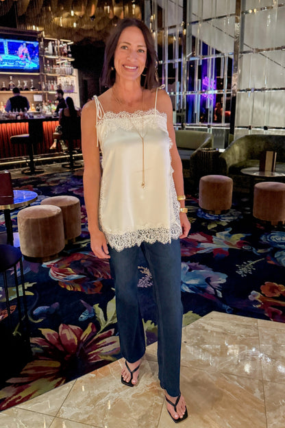 Front view of woman wearing Swagger Boutique’s Ivory Satin Lace Trim Tank Top by Entro with jeans standing at a restaurant and bar.