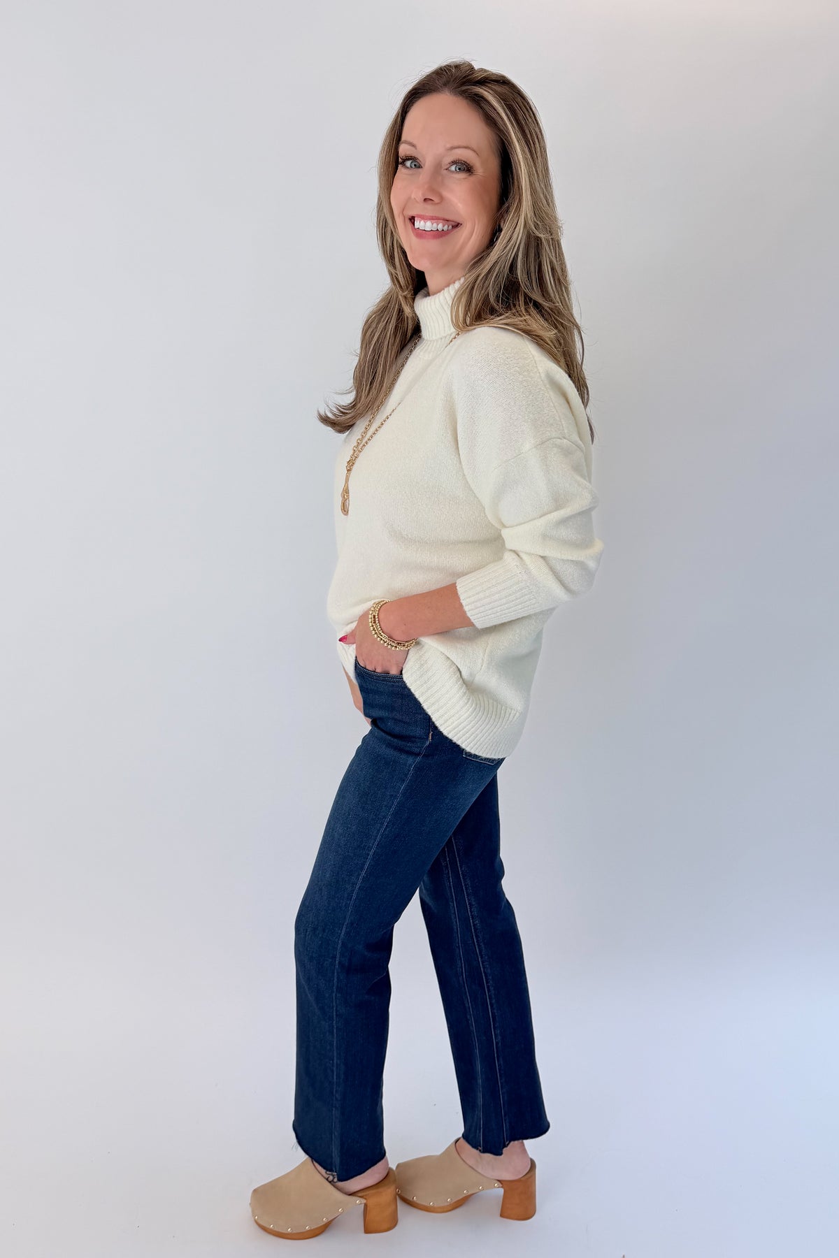 Side view of woman wearing Swagger Boutique’s Ivory Turtleneck Sweater with jeans against white studio background.