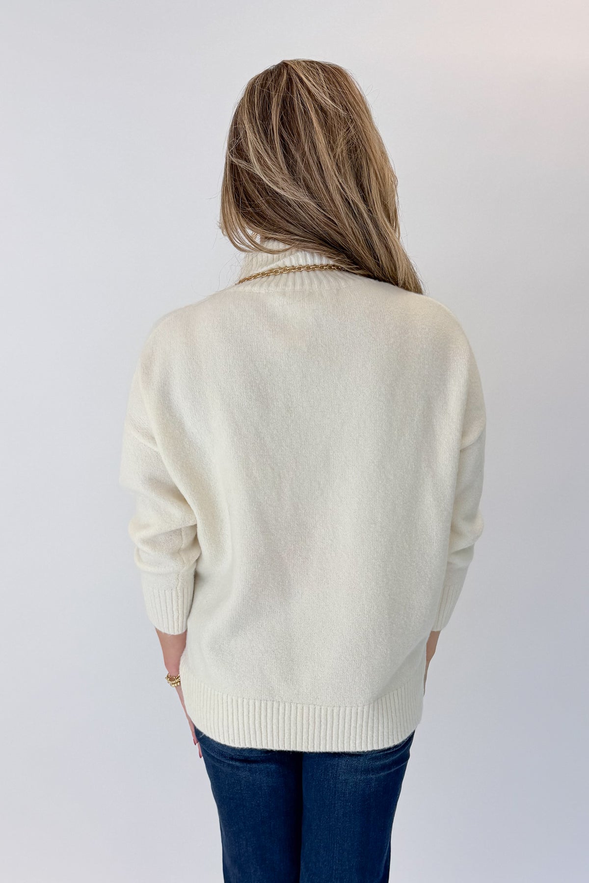 Back view of woman wearing Swagger Boutique’s Ivory Turtleneck Sweater with jeans against white studio background.
