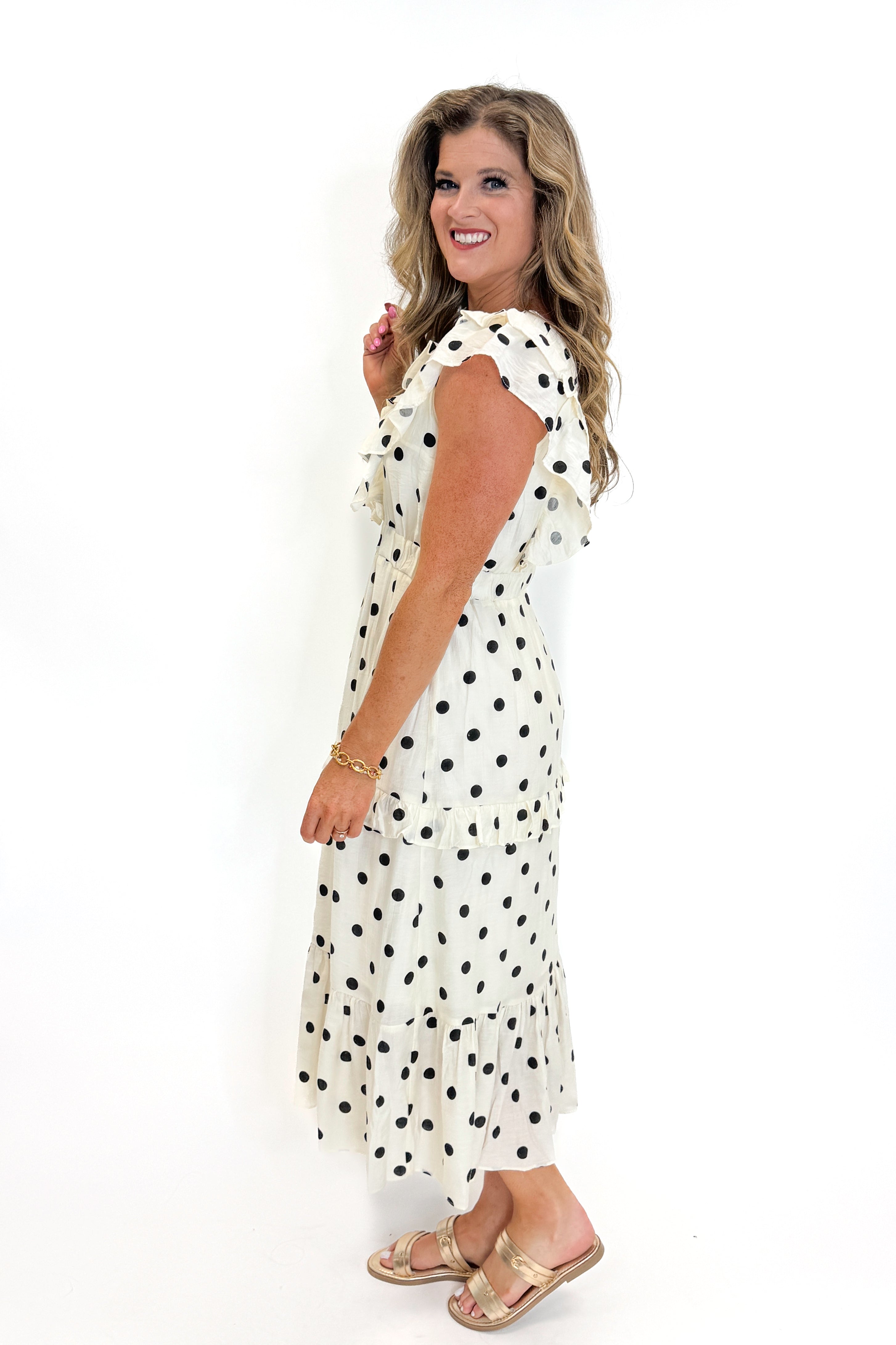 Side view of woman wearing Swagger Boutique’s Ivory &amp; Black Polka Dot Ruffle Dress with sandals against white studio background.