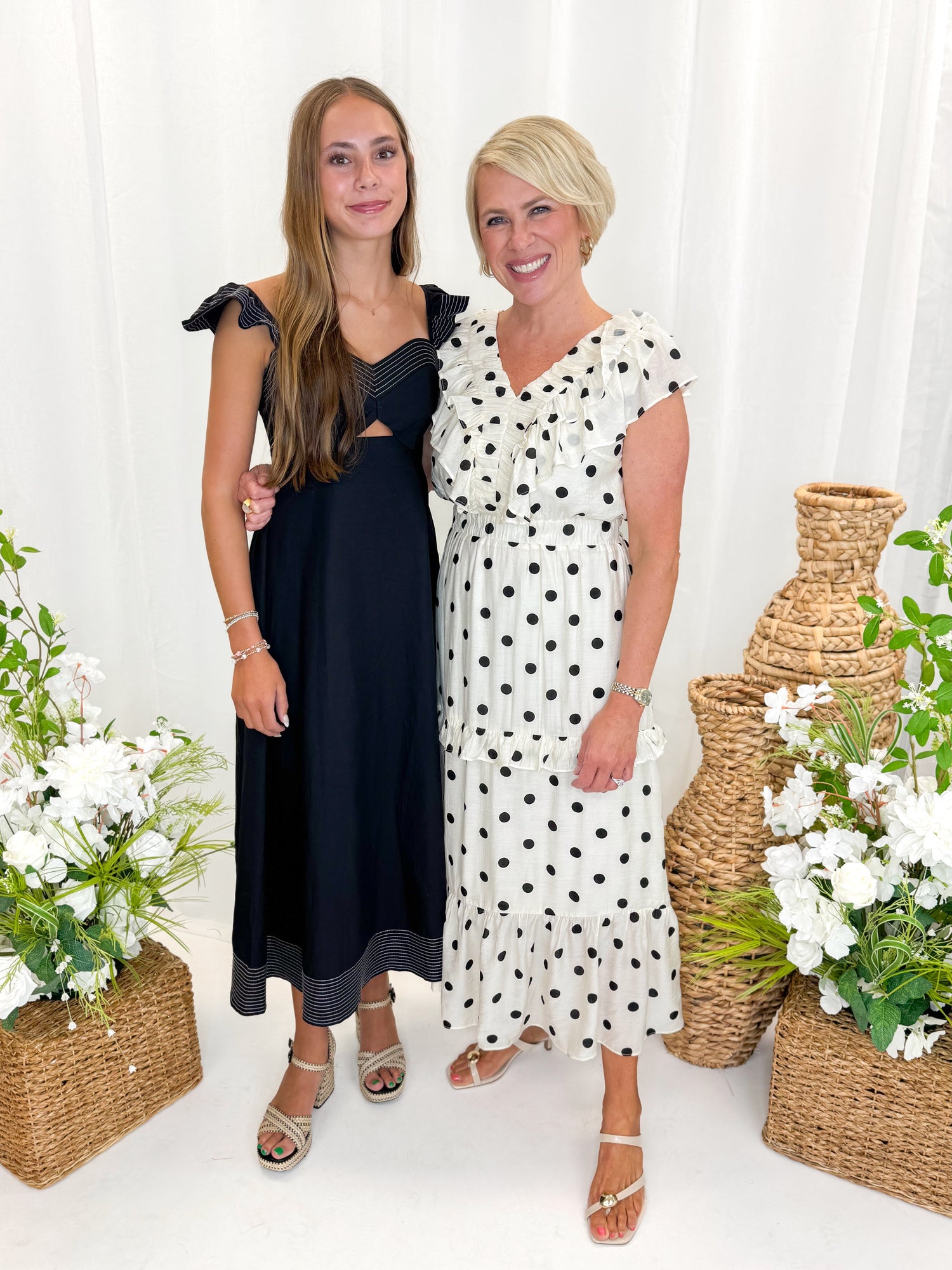 Front view of woman wearing Swagger Boutique’s Ivory &amp; Black Polka Dot Ruffle Dress with sandals and daughter against decorative background.