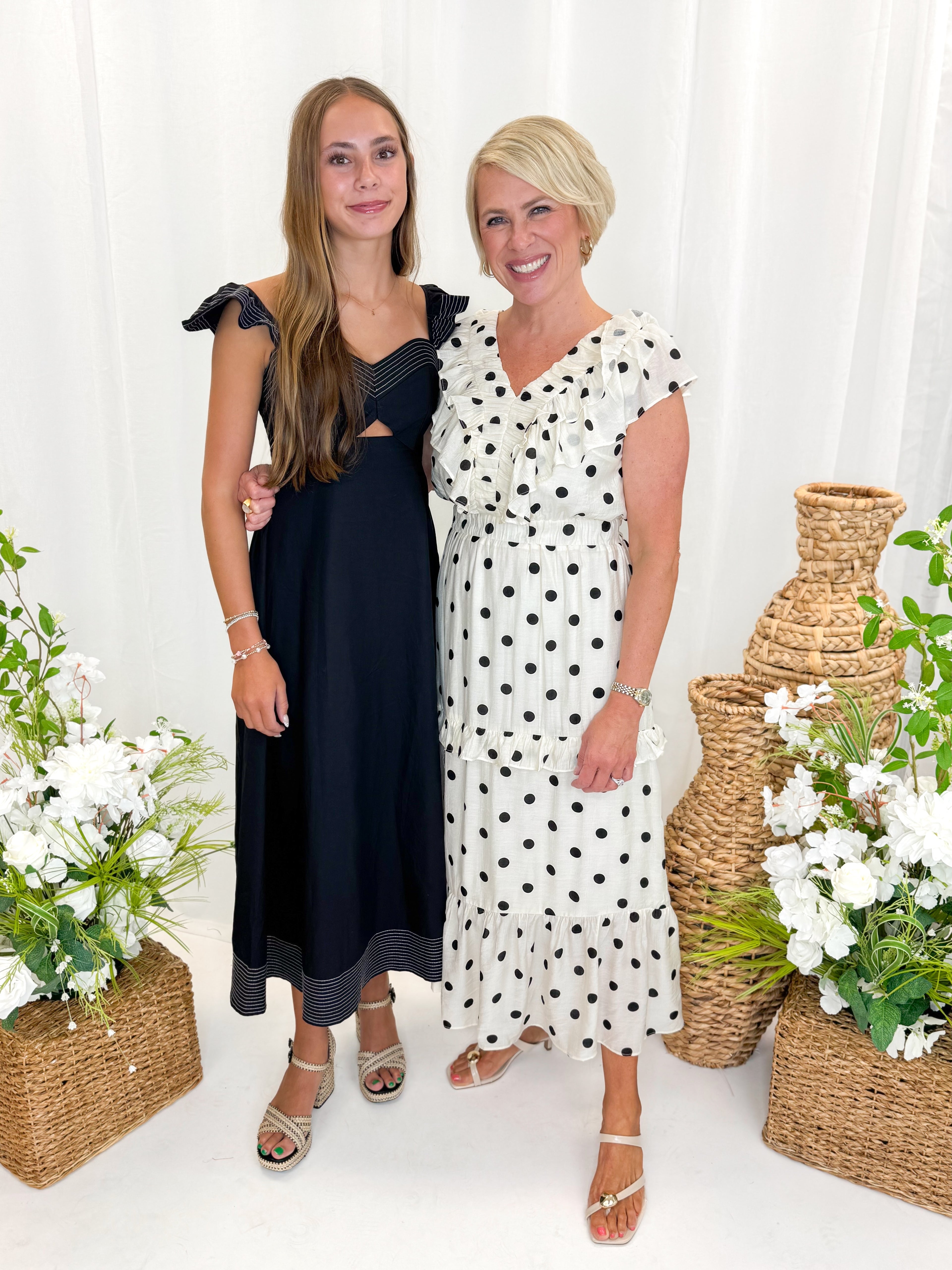 Front view of woman wearing Swagger Boutique’s Ivory &amp; Black Polka Dot Ruffle Dress with sandals and daughter against decorative background.