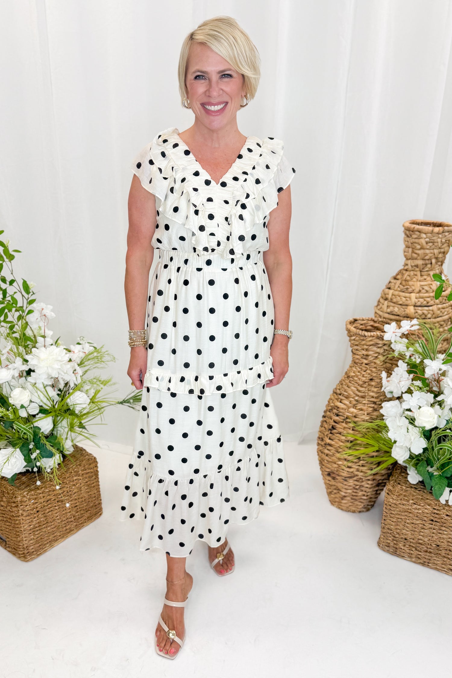 Front view of woman wearing Swagger Boutique’s Ivory &amp; Black Polka Dot Ruffle Dress with sandals against decorative background.