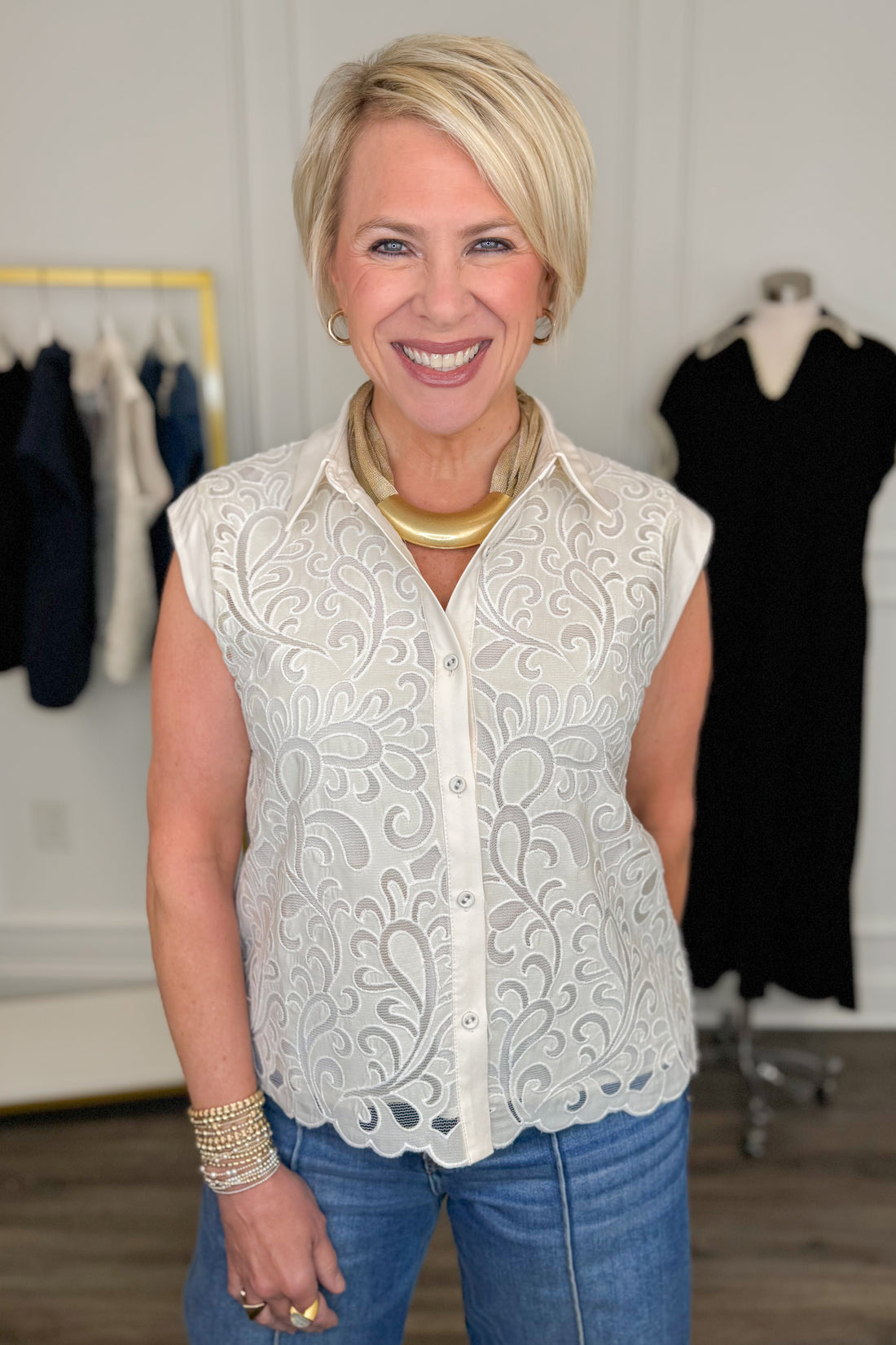 Front view of woman wearing Swagger Boutique’s Ivory Lace Sleeveless Top with jeans with clothing hanging in the background.