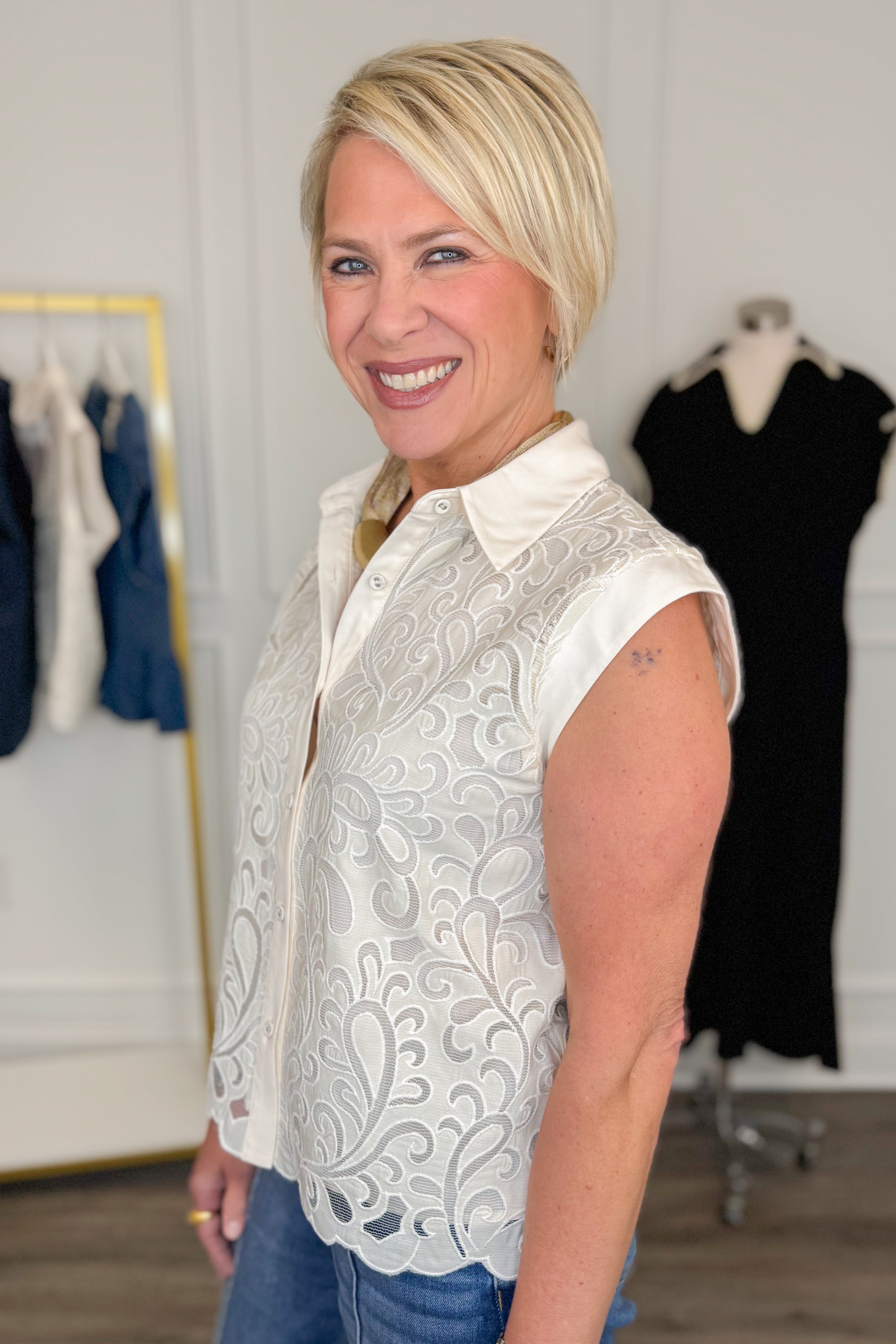 Side view of woman wearing Swagger Boutique’s Ivory Lace Sleeveless Top with jeans with clothing hanging in the background.
