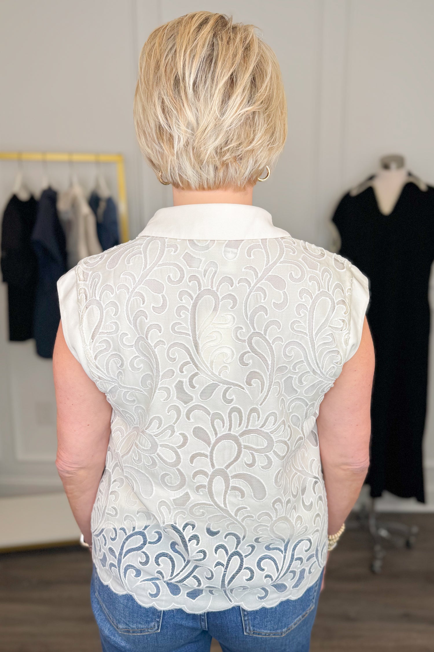 Back view of woman wearing Swagger Boutique’s Ivory Lace Sleeveless Top with jeans with clothing hanging in the background.