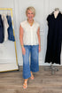 Front view of woman wearing Swagger Boutique’s Ivory Lace Sleeveless Top with jeans with clothing hanging in the background.