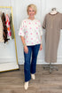 Front view of woman wearing Swagger Boutique’s Ivory & Pink Polka Dot Short Sleeve Cardigan with jeans with clothing hanging in the background.