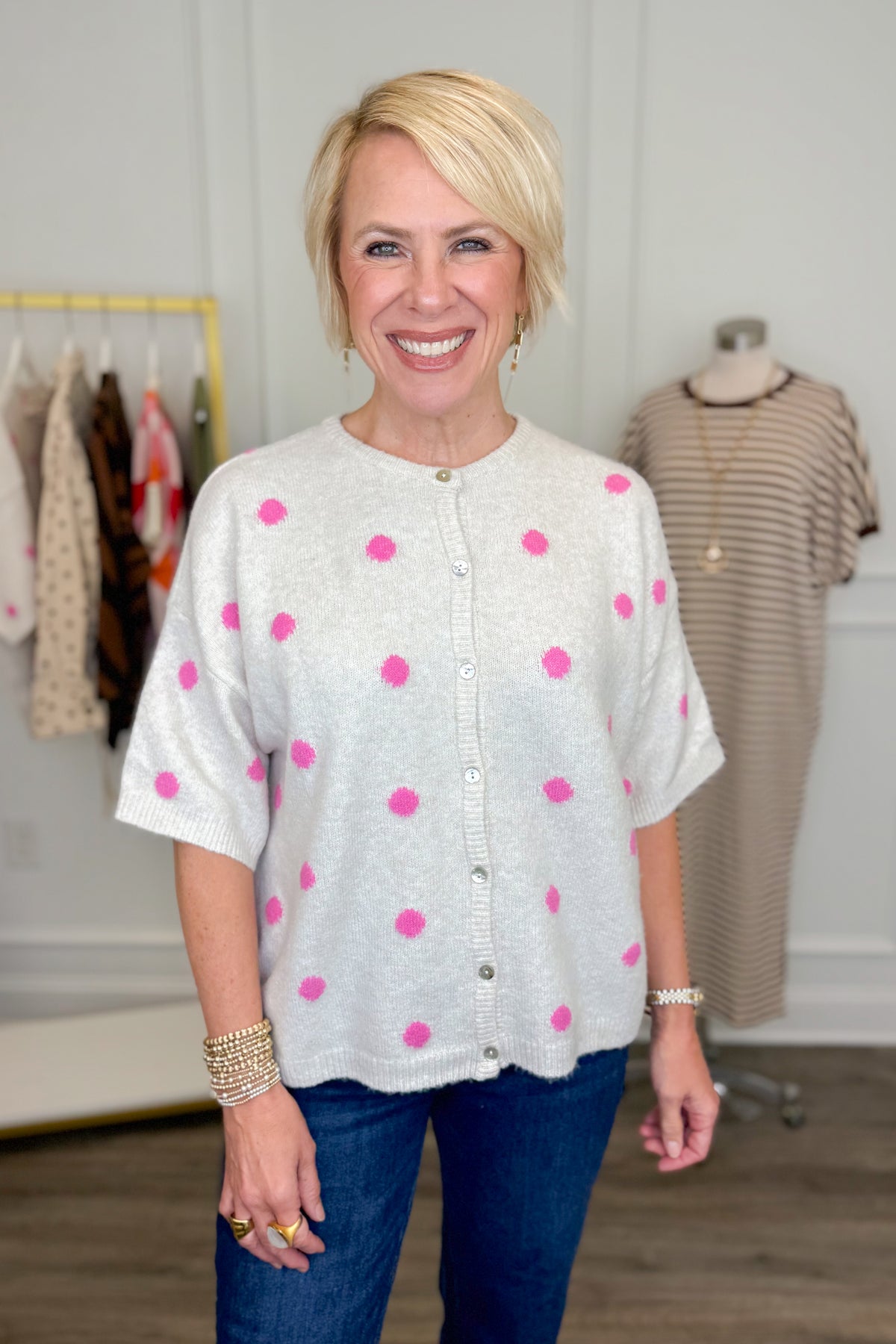 Front view of woman wearing Swagger Boutique’s Ivory &amp; Pink Polka Dot Short Sleeve Cardigan with jeans with clothing hanging in the background.