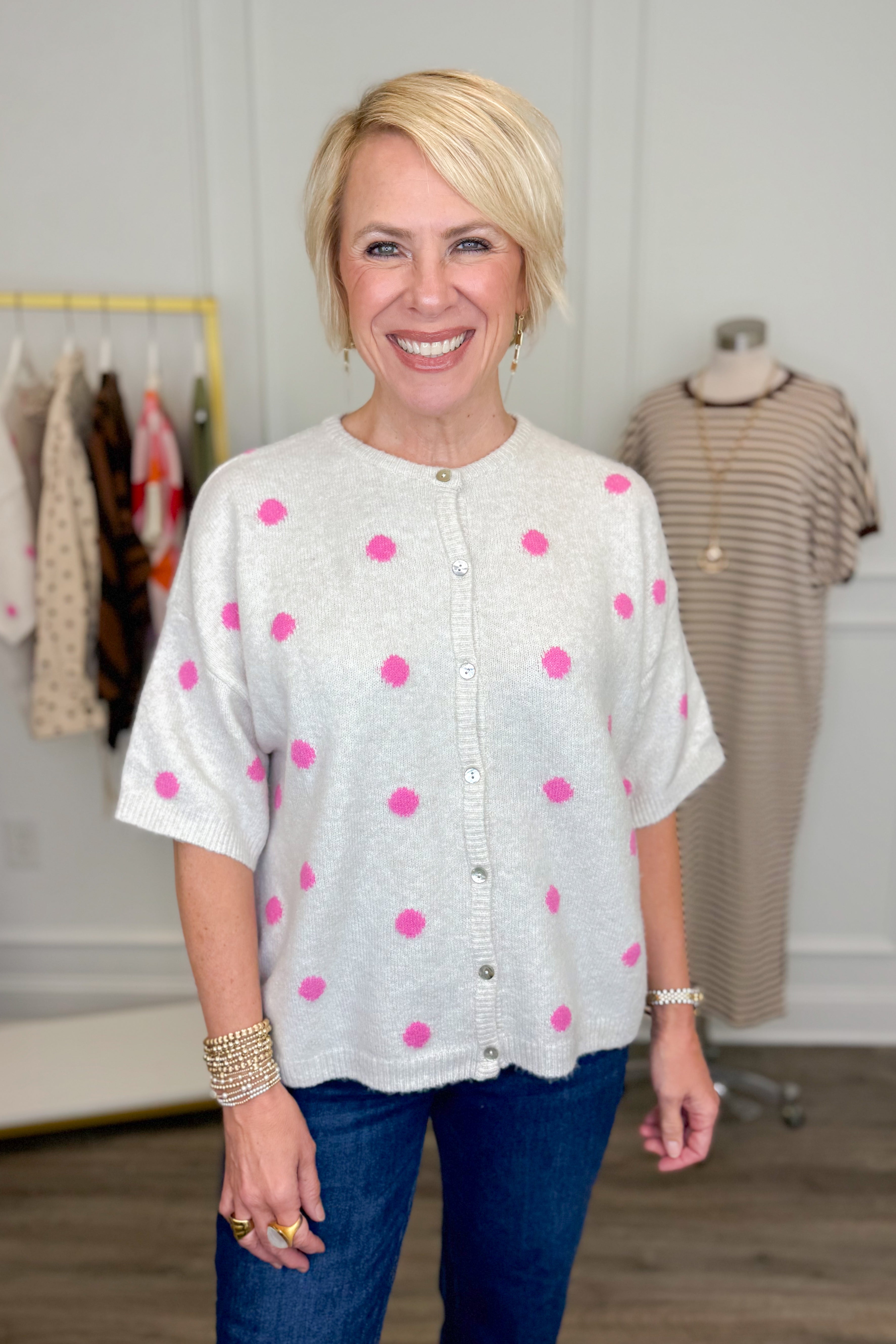 Front view of woman wearing Swagger Boutique’s Ivory &amp; Pink Polka Dot Short Sleeve Cardigan with jeans with clothing hanging in the background.