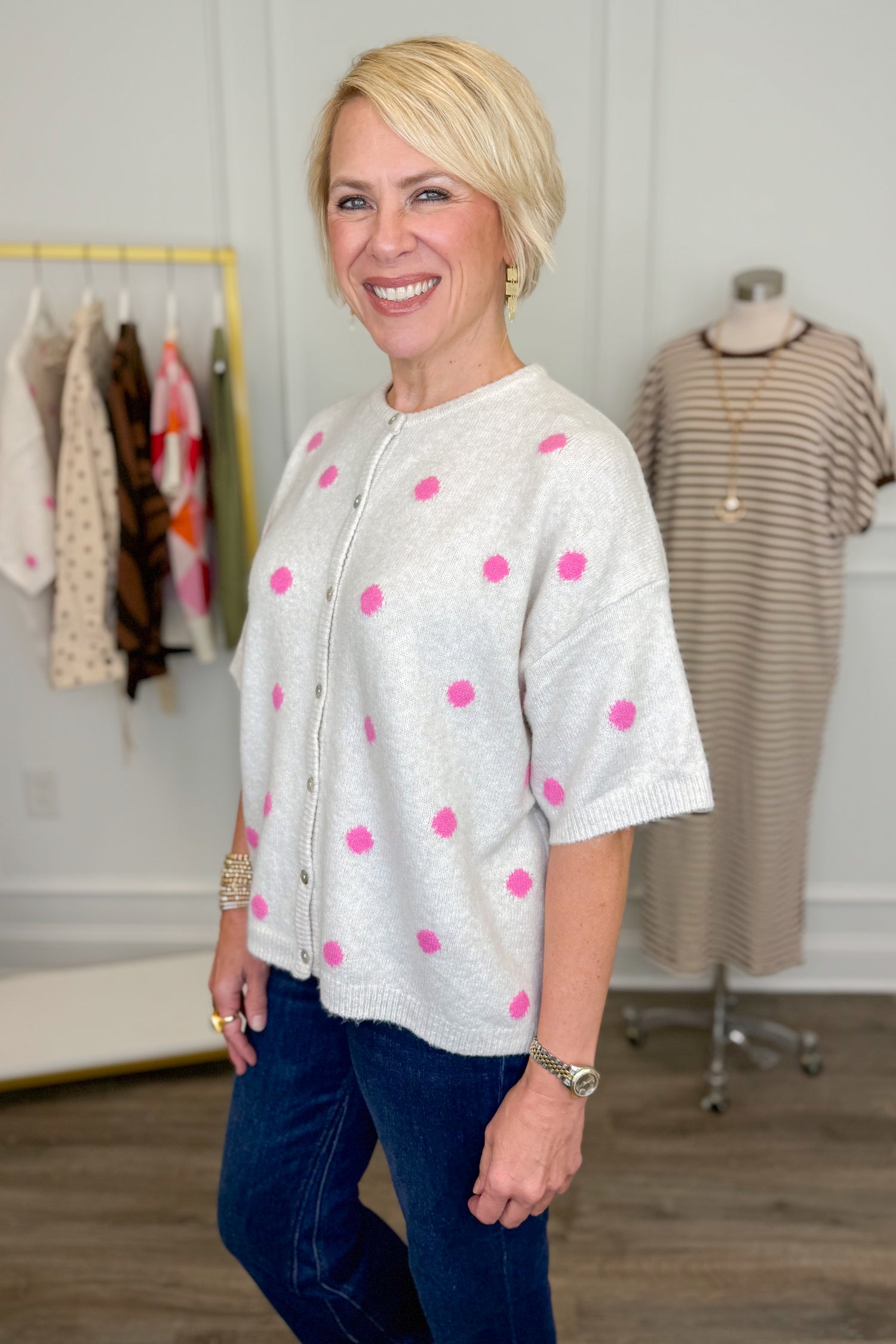 Side view of woman wearing Swagger Boutique’s Ivory &amp; Pink Polka Dot Short Sleeve Cardigan with jeans with clothing hanging in the background.