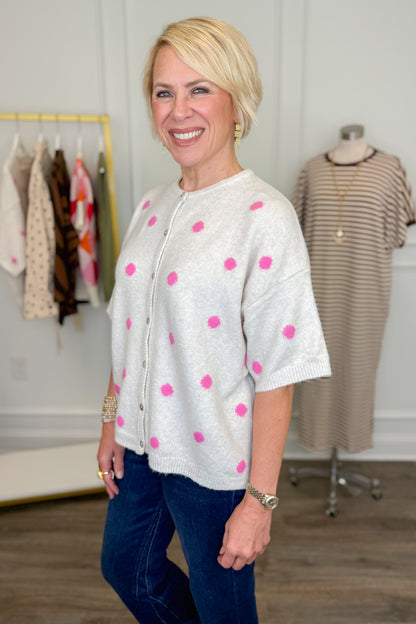 Side view of woman wearing Swagger Boutique’s Ivory &amp; Pink Polka Dot Short Sleeve Cardigan with jeans with clothing hanging in the background.