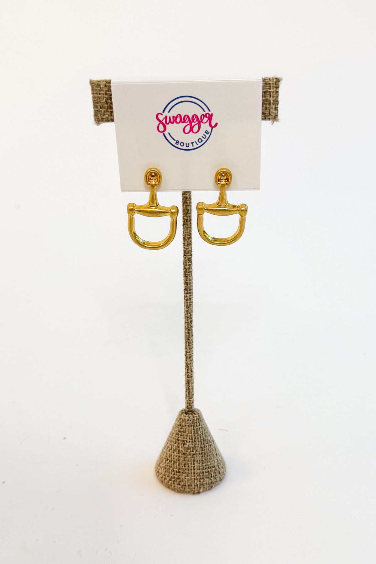 A pair of gold horsebit stud earrings displayed on a burlap-covered stand with a white card labeled Swagger Boutique