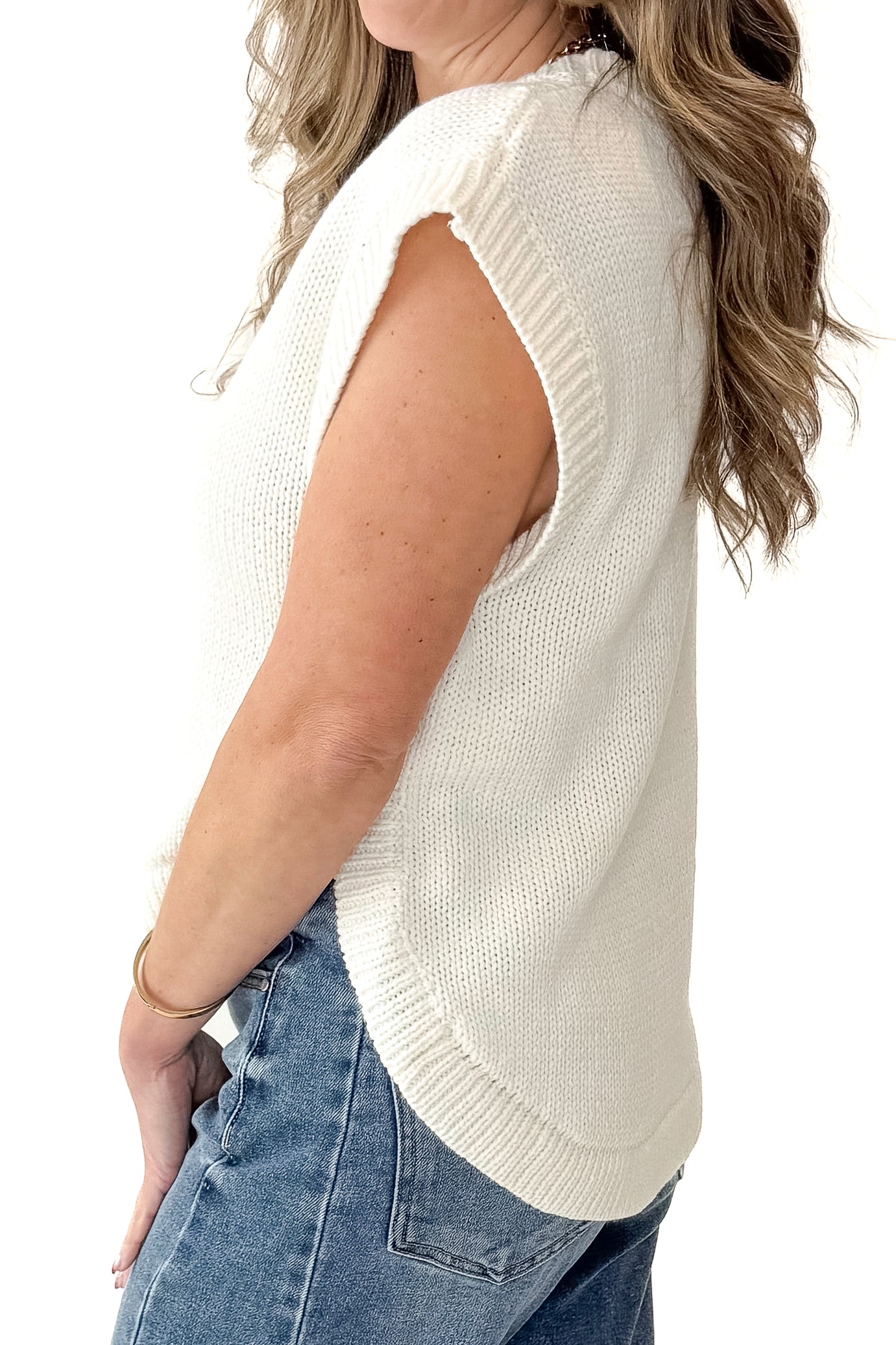 Side view of woman wearing Cream Side Button Detail Sleeveless Sweater with jeans against white studio background.