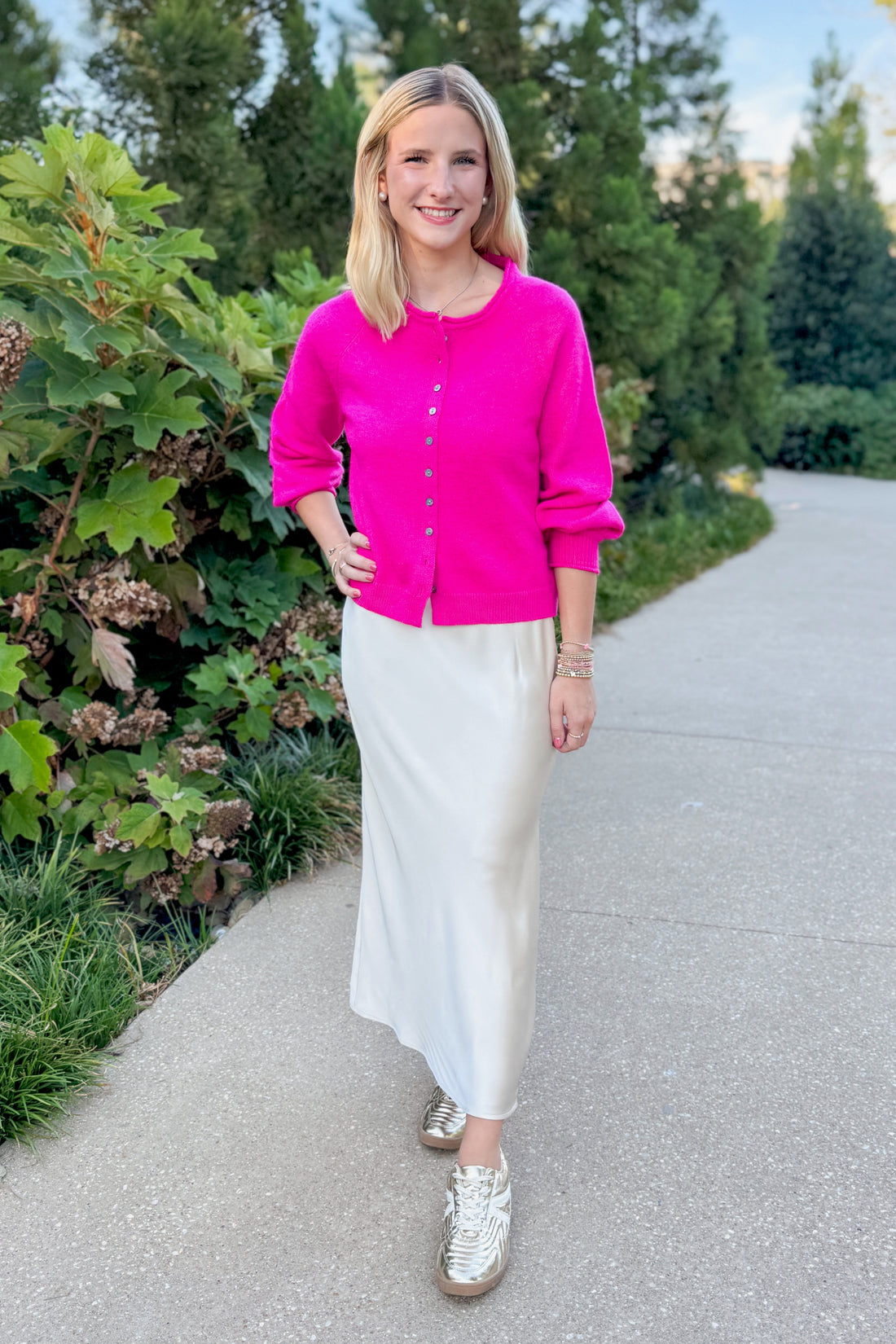 Woman in a pink cardigan and white skirt standing on a path with greenery.