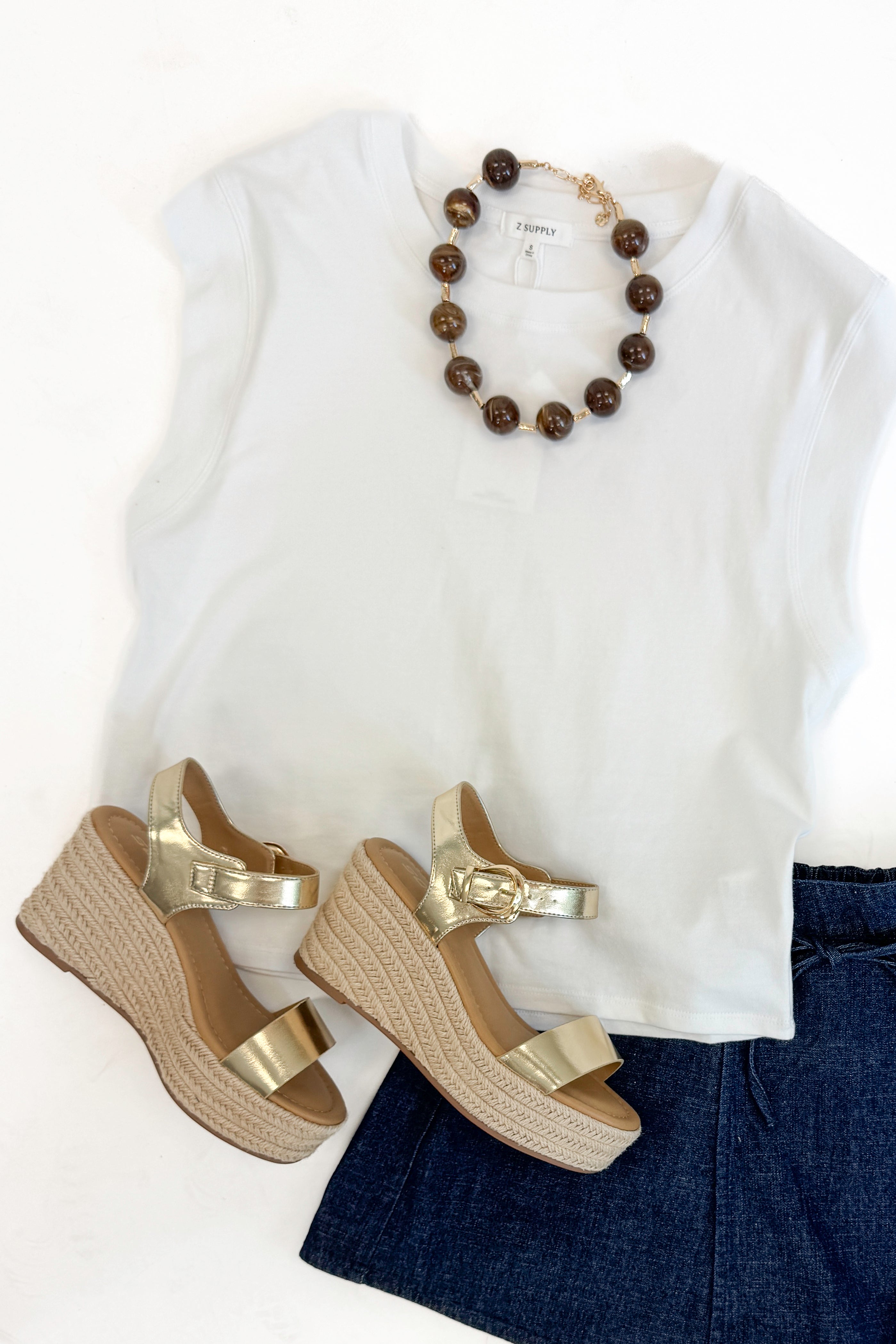 Flat lay with white top, jean shorts, chunky necklace and Jena Gold and Rattan Wedge shoes against white studio background.