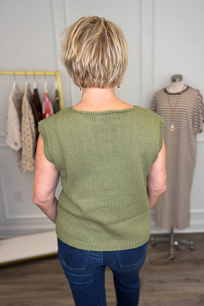 A woman viewed from behind wearing a textured sage green sleeveless top and blue jeans in a boutique setting.