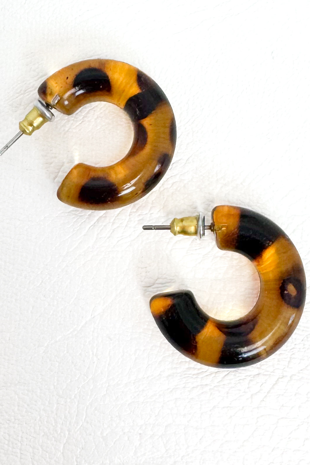 A pair of chunky C-shaped hoop earrings with a glossy tortoiseshell pattern in amber brown, black, and tan, featuring gold-tone posts on a textured white background.