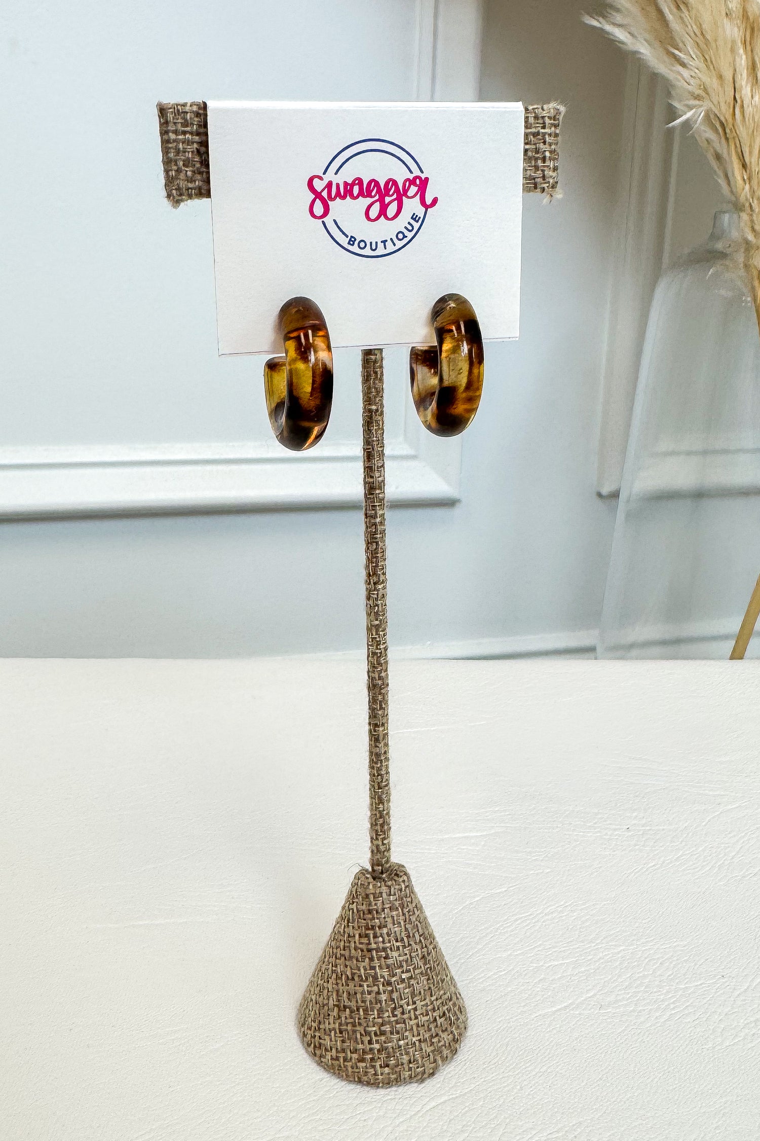 A pair of large gold hoop earrings displayed on a white card stand with burlap trim, set on a white surface against a light gray wall background.