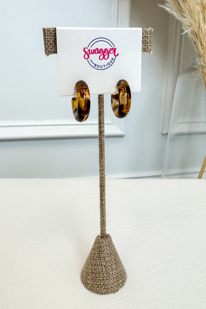 A pair of large gold hoop earrings displayed on a white card stand with burlap trim, set on a white surface against a light gray wall background.