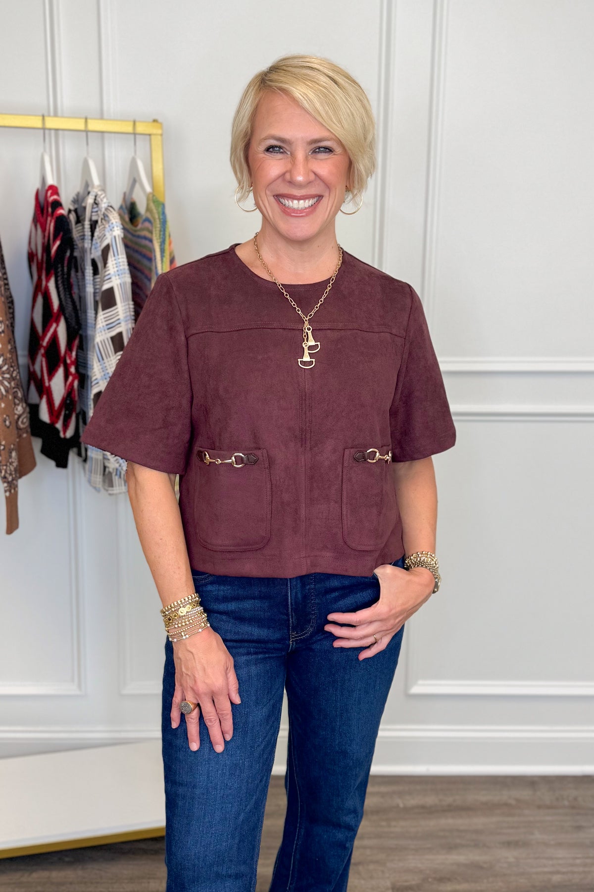 A smiling blonde woman wearing a short-sleeve dusty rose-brown faux suede cropped top with gold-tone hardware and front pockets, paired with blue jeans, standing in a boutique setting.