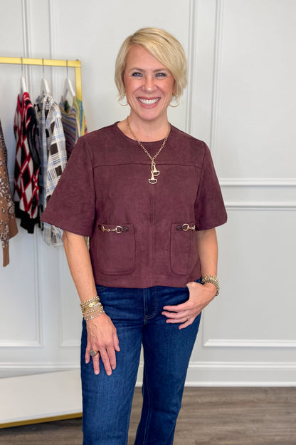 A smiling blonde woman wearing a short-sleeve dusty rose-brown faux suede cropped top with gold-tone hardware and front pockets, paired with blue jeans, standing in a boutique setting.