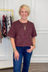 A smiling blonde woman wearing a short-sleeve dusty rose-brown faux suede cropped top with gold-tone hardware and front pockets, paired with blue jeans, standing in a boutique setting.
