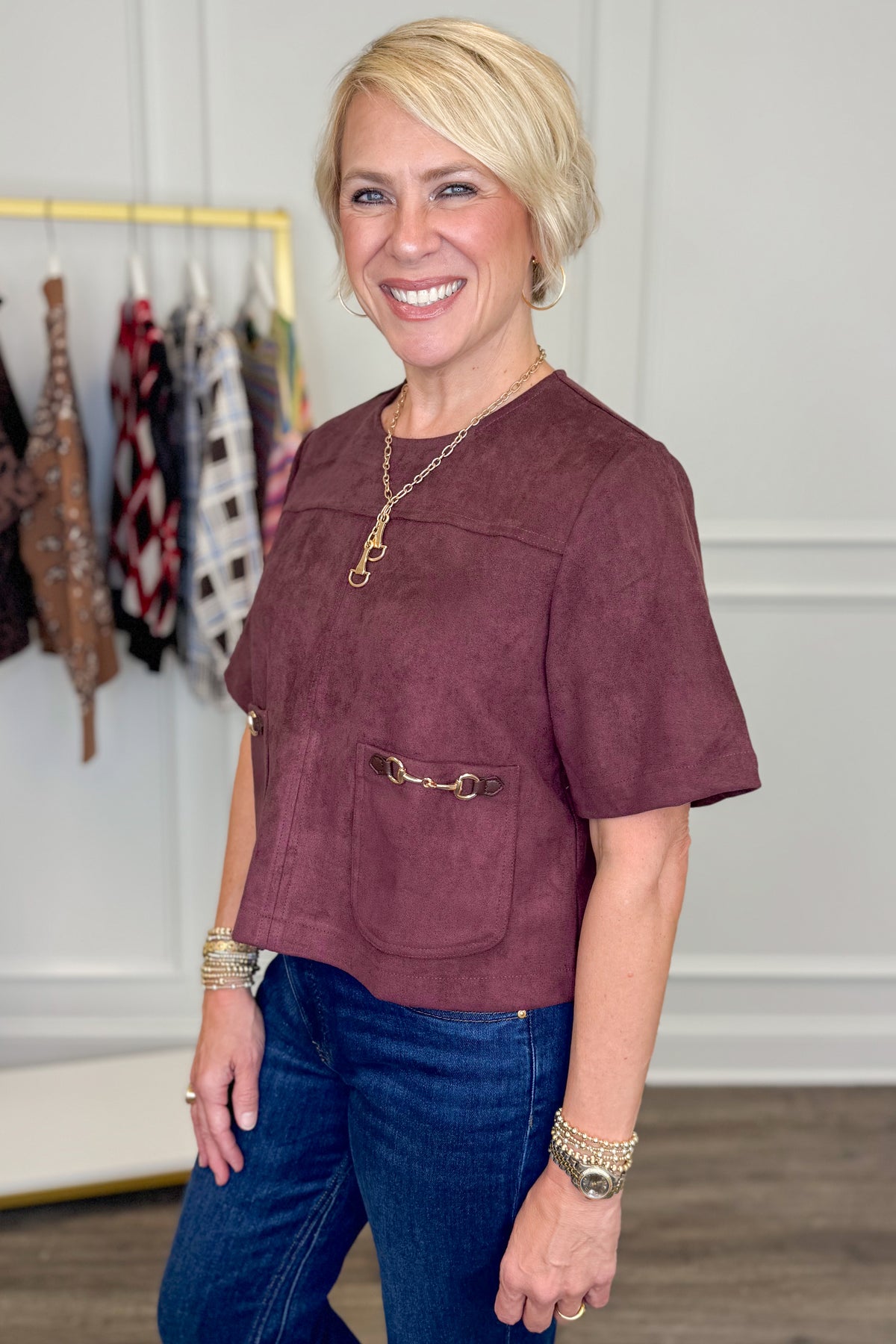 A smiling blonde woman wearing a short-sleeve dusty burgundy top with a front patch pocket and gold-tone hardware, paired with blue jeans, standing in a boutique interior.