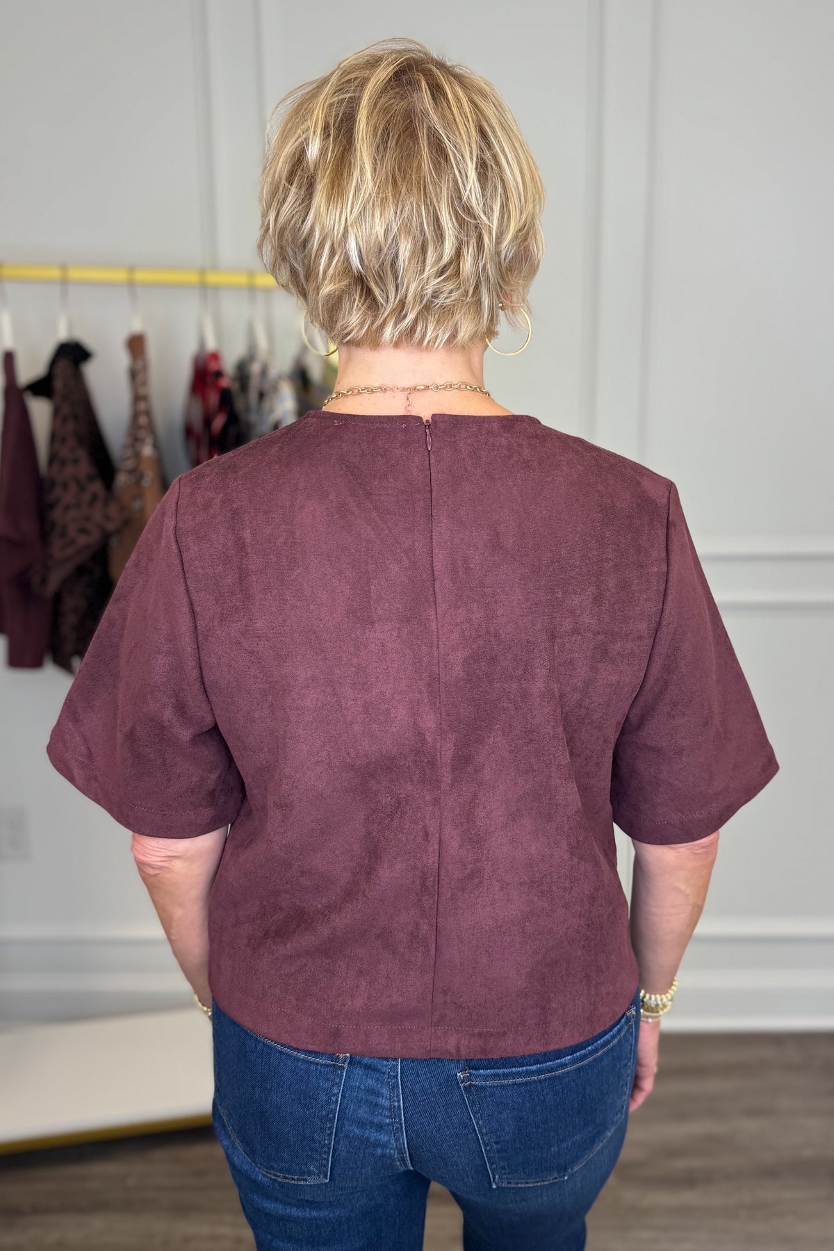 A woman viewed from behind wearing a short-sleeve dusty plum-brown top and blue jeans in an indoor boutique setting.