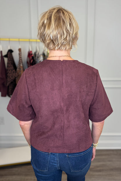 A woman viewed from behind wearing a short-sleeve dusty plum-brown top and blue jeans in an indoor boutique setting.