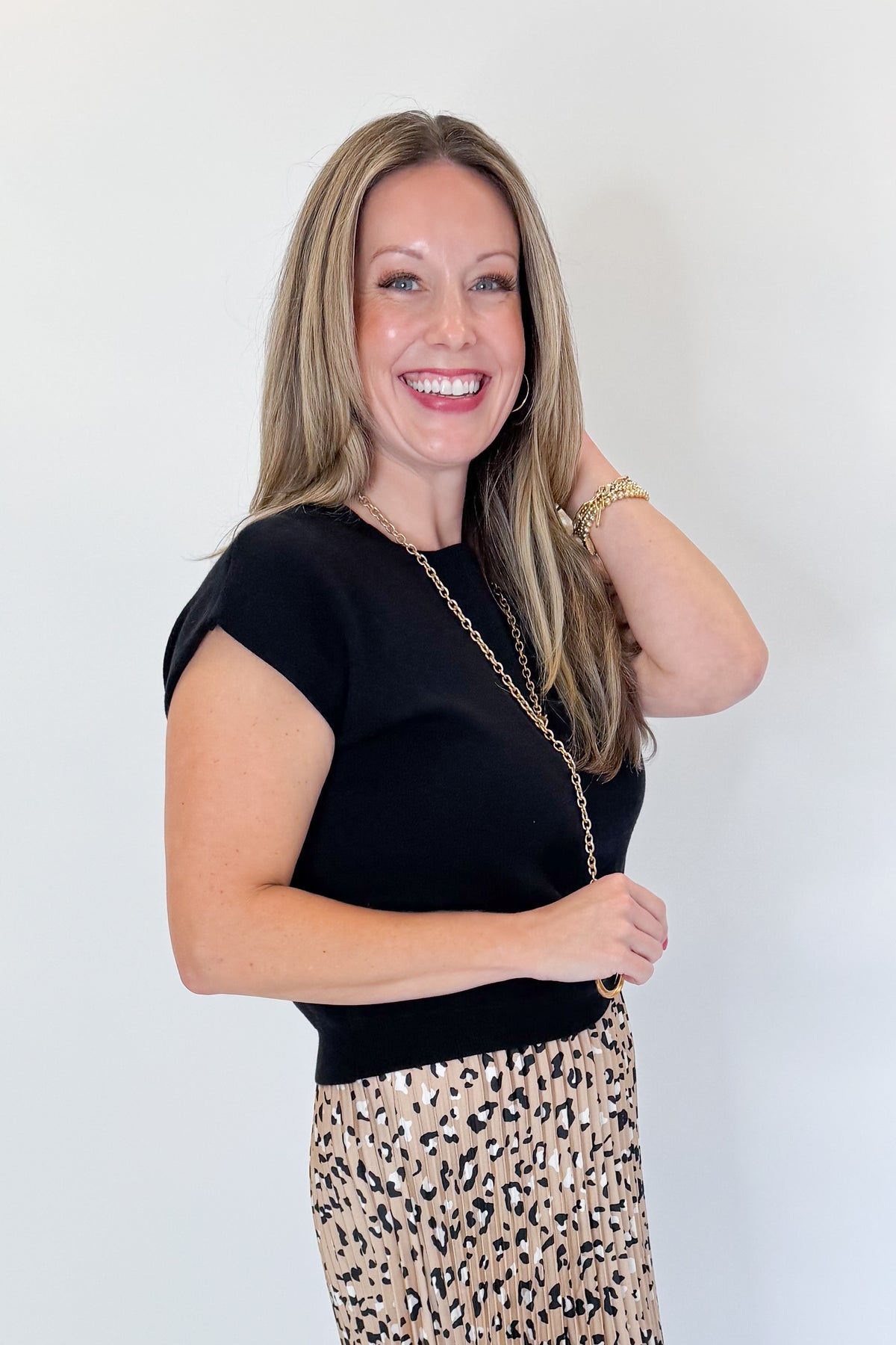 Side view of woman wearing Swagger Boutique’s Leopard Pleated Skirt &amp; Black Short Sleeve Twist Detail Sweater against white studio background.