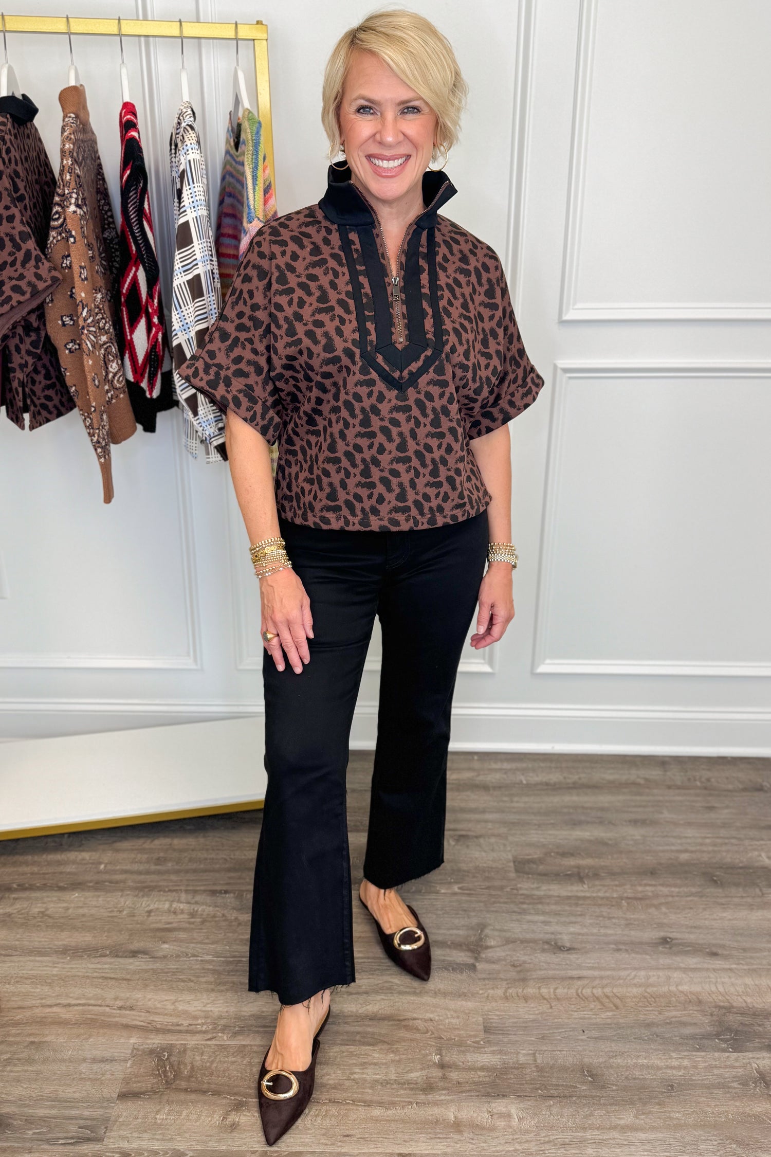 Front view of woman wearing Swagger Boutique’s Leopard Half Zip Top by Entro with jeans with clothing hanging in the background.