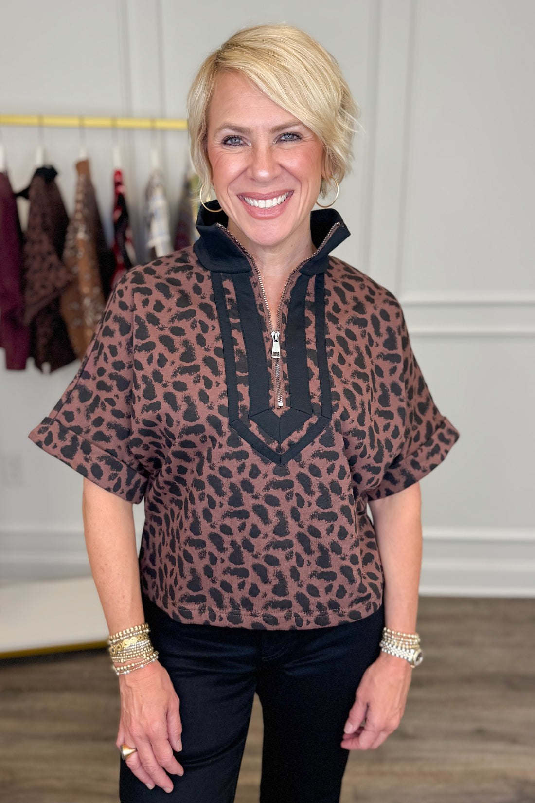 Front view of woman wearing Swagger Boutique’s Leopard Half Zip Top by Entro with jeans with clothing hanging in the background.