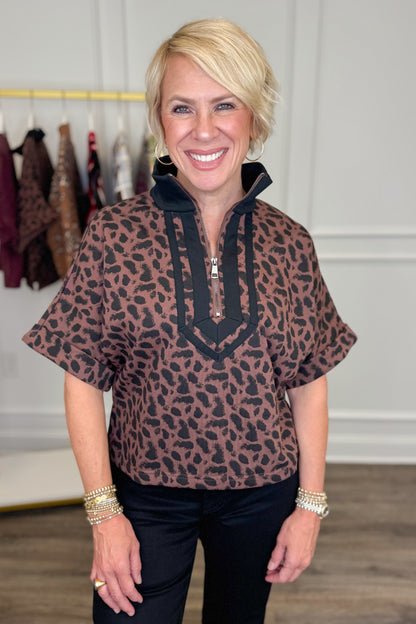 Front view of woman wearing Swagger Boutique’s Leopard Half Zip Top by Entro with jeans with clothing hanging in the background.