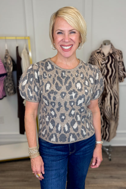 Front view of woman wearing Swagger Boutique’s Leopard Short Sleeve Sweater with jeans with clothing hanging in the background.