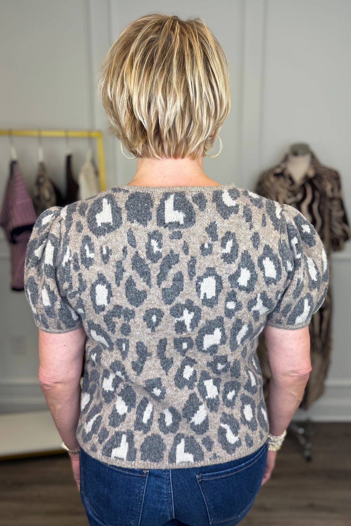 Back view of woman wearing Swagger Boutique’s Leopard Short Sleeve Sweater with jeans with clothing hanging in the background.