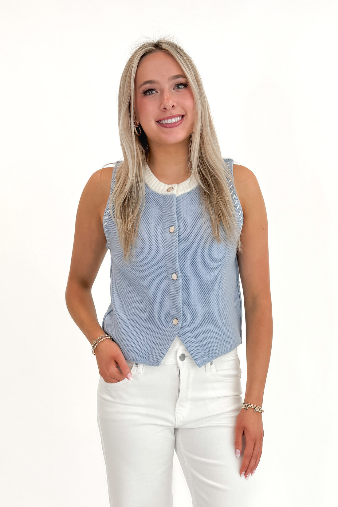 Front view of woman wearing Light Blue Button Up Sleeveless Sweater with white jeans against white studio background.