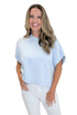 A smiling woman with blonde hair wearing a light blue short-sleeve knit top and white pants, standing against a plain white background.