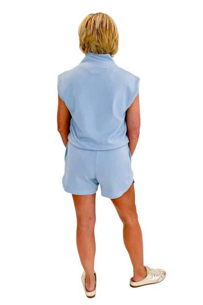 Back view of woman wearing Swagger Boutique’s Light Blue Sleeveless 1/4 Zip Top with matching shorts against white studio background.