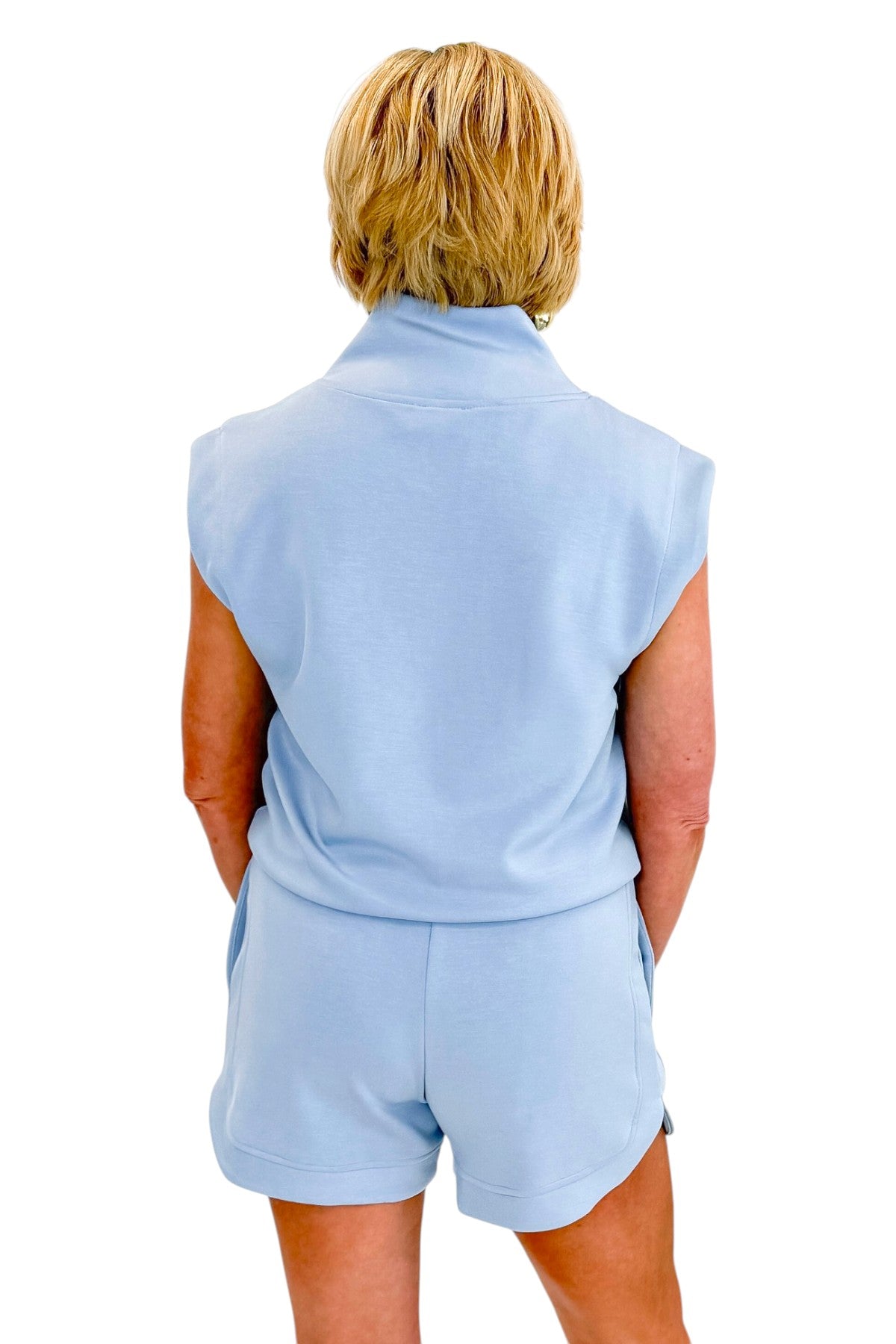 Back view of woman wearing Swagger Boutique’s Light Blue Sleeveless 1/4 Zip Top with matching shorts against white studio background.
