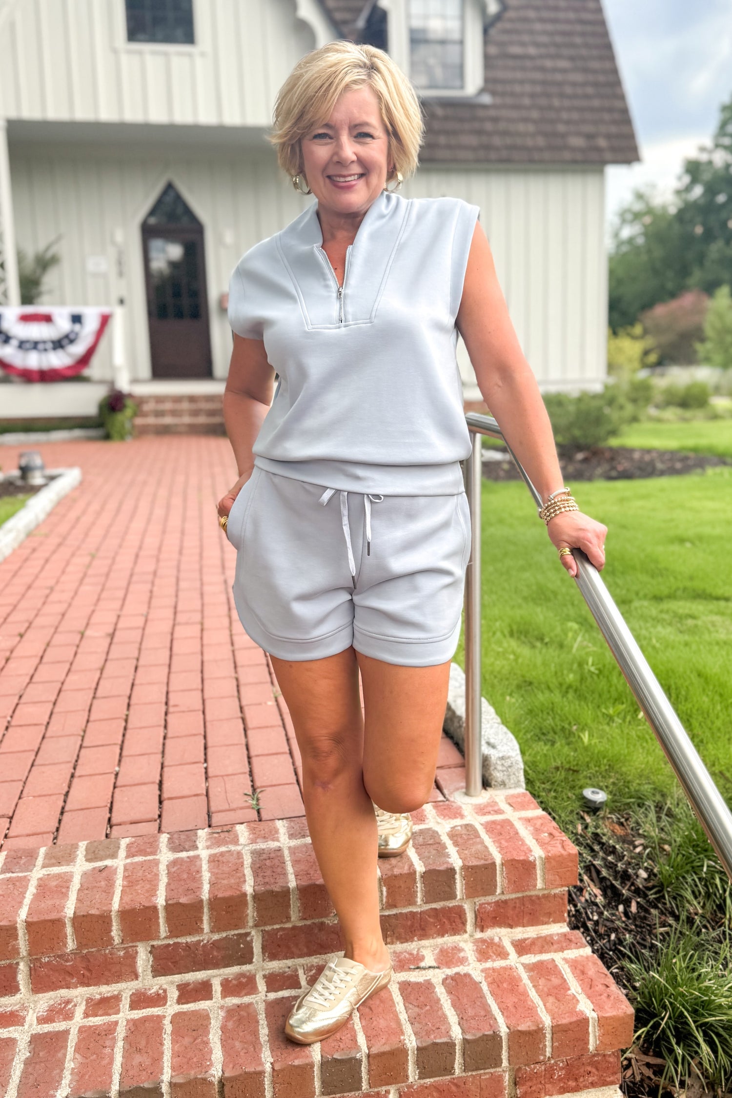 Front view of woman wearing Swagger Boutique’s Light Blue Sleeveless 1/4 Zip Top with matching shorts walking down a brick path outside.