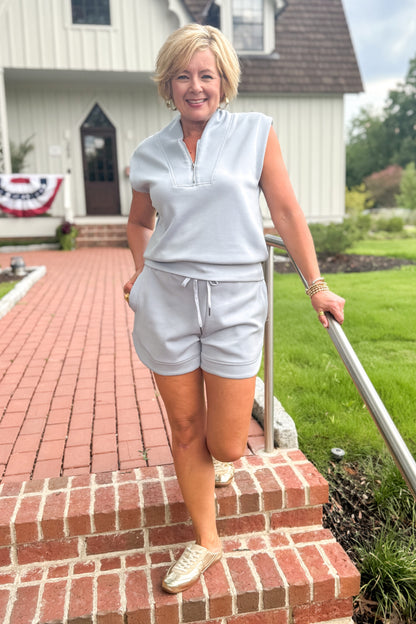 Front view of woman wearing Swagger Boutique’s Light Blue Sleeveless 1/4 Zip Top with matching shorts walking down a brick path outside.
